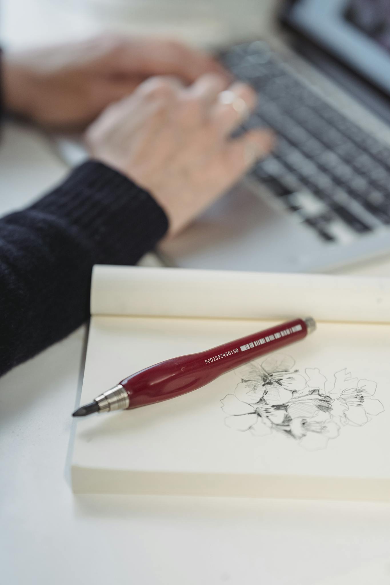 Close-up of a pencil and flower sketches on a notebook with hands typing on a laptop.